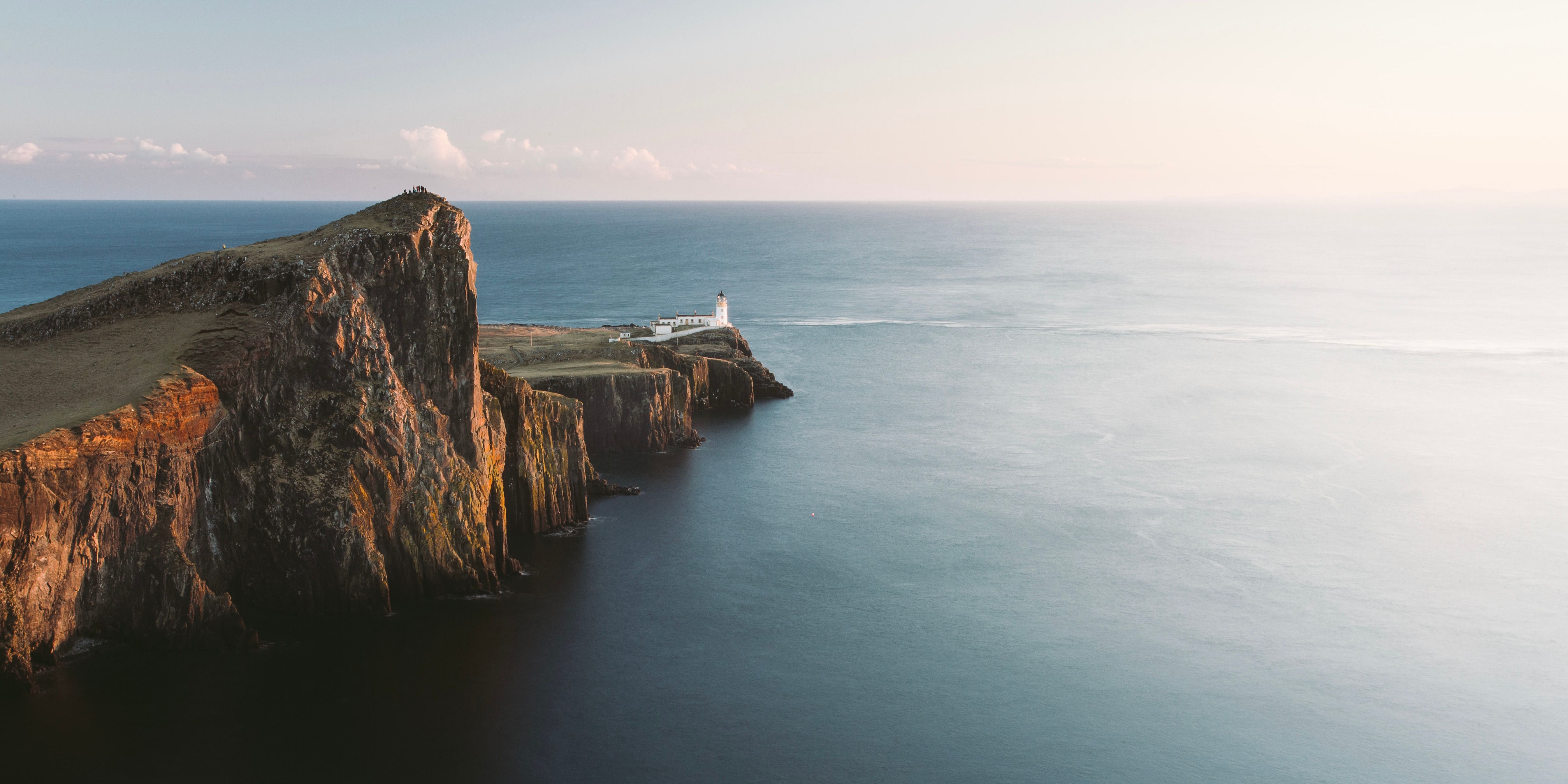 A breathtaking view of the Neist Point Lighthouse perched on the rugged cliffs of the Isle of Skye, Scotland, overlooking the calm sea at sunset.